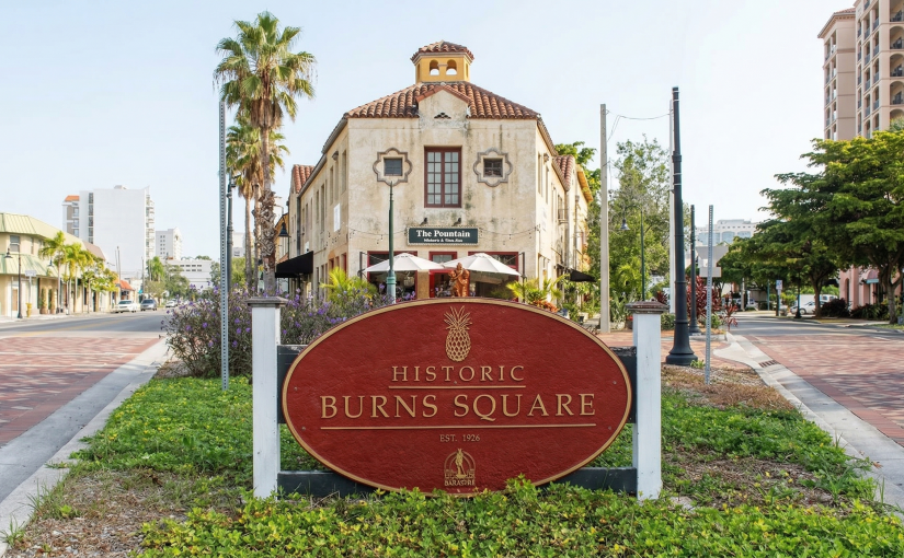 A prominent, oval, red and gold sign centered in the foreground in a landscaped median, reading "HISTORIC BURNS SQUARE," "EST. 1926," with a pineapple crest. Behind it is a brick-paved intersection with a Mediterranean-style building (labeled "The Fountain") and tall palm trees under a blue sky, leading into a walkable street scene.