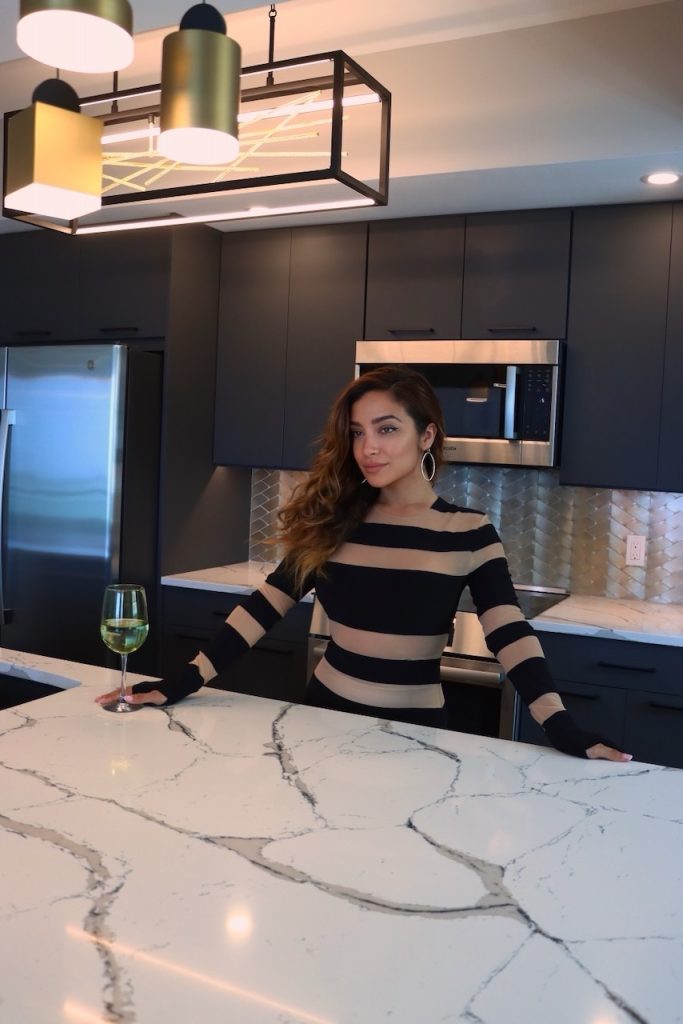 A woman in a black and tan striped dress holding a glass of white wine behind a large white marble kitchen island in a modern dark kitchen.