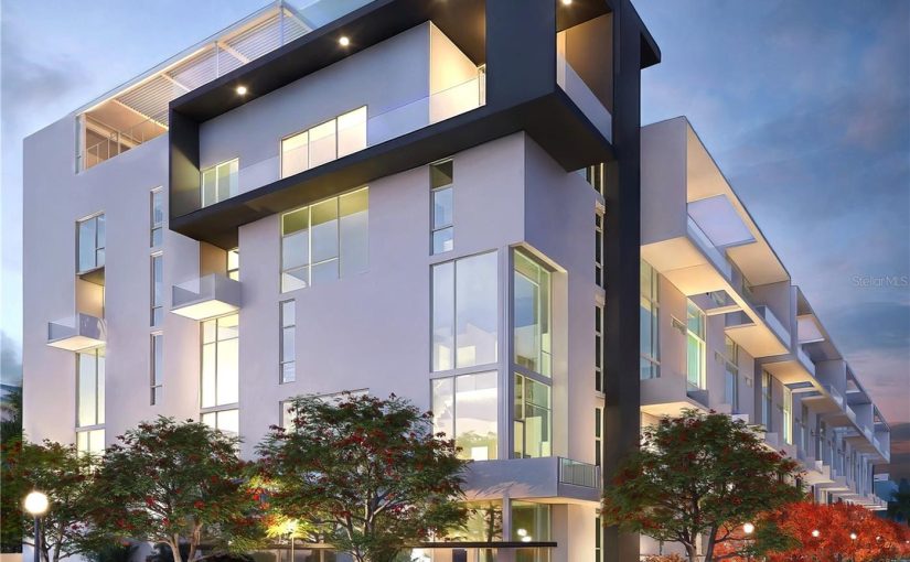 A modern, multi-story luxury condominium building featuring white and dark gray architectural accents, floor-to-ceiling glass windows, and landscaped sidewalks at twilight.