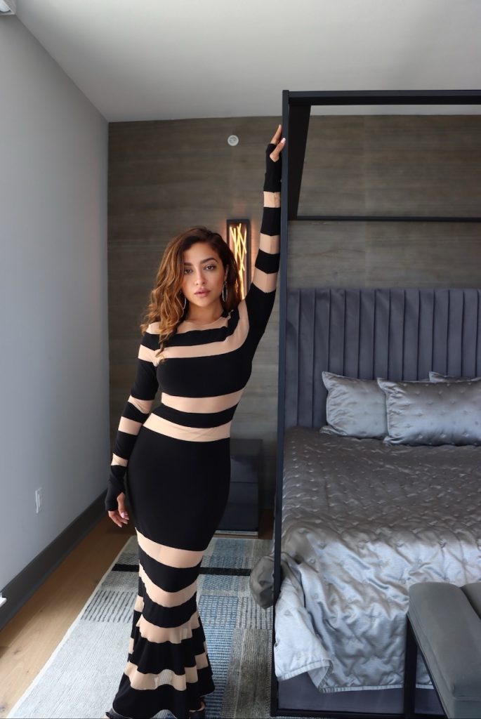 A woman leaning against a black canopy bed frame in a bedroom with a wood-paneled accent wall and grey tufted headboard.