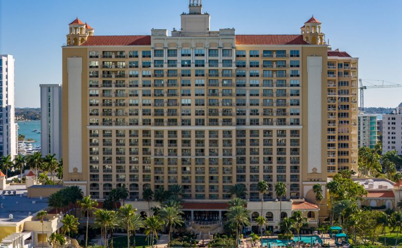 Front exterior view of the multi-story Ritz-Carlton Hotel in downtown Sarasota, featuring Mediterranean-style architecture, a waterfront swimming pool, and palm trees under a clear blue sky.