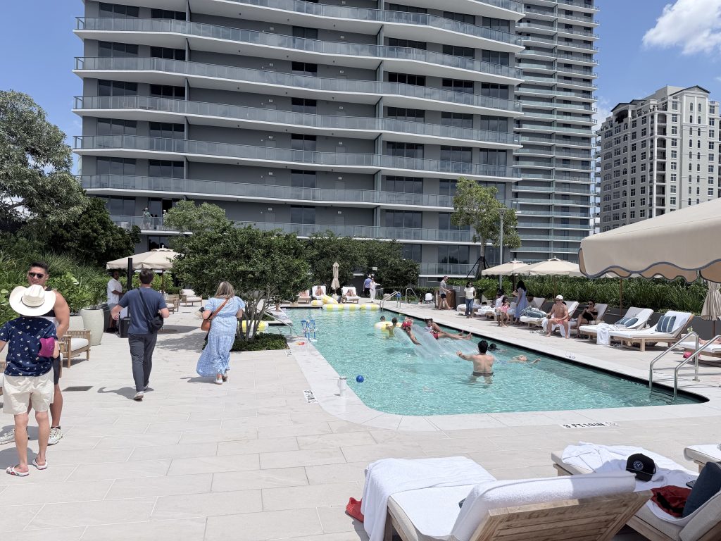 Resort-style swimming pool with residents and guests at the Ritz-Carlton Residences, Tampa by Related. Modern high-rise tower with expansive balconies overlooks the lively pool area on a sunny day.