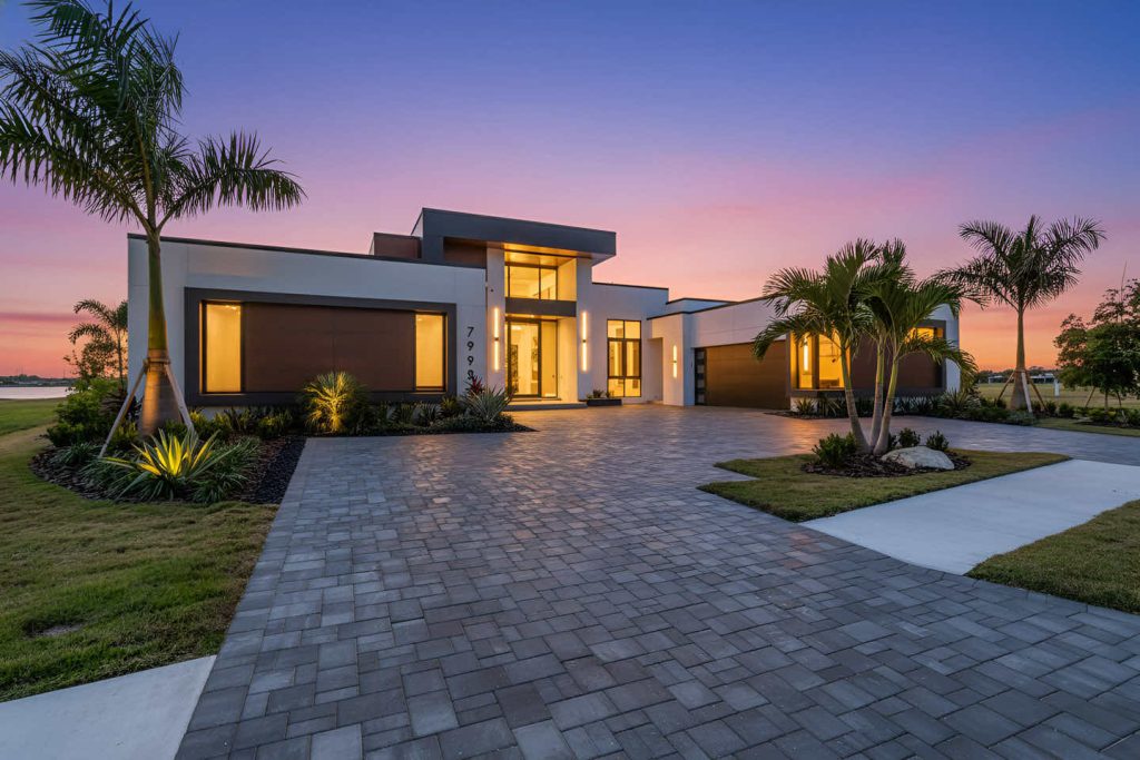 Modern house with a paved driveway and palm trees under a pink and orange sunset.